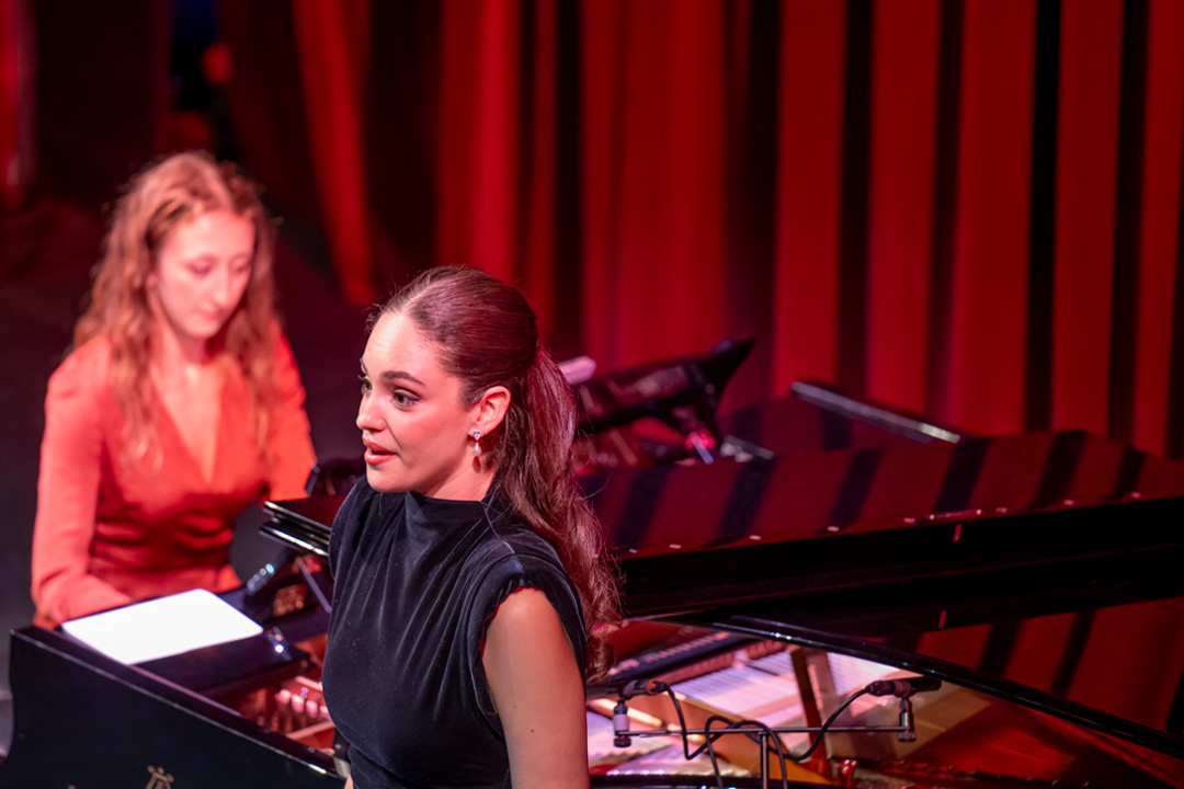 A women wearing a black dress, singing on stage, with a piano behind her with a pianist accompanying her on stage, with a red curtain.